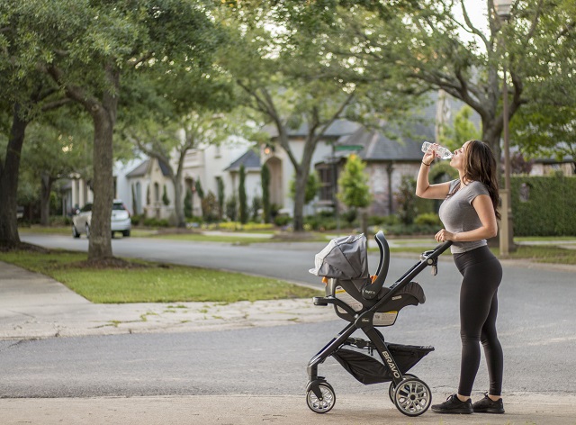 picture of a woman drinking water while pushing baby in a stroller on a road 